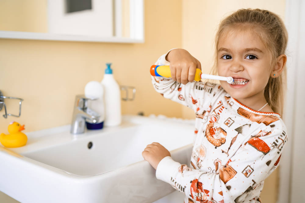 girl brushing her teeth with an electric brush, standing in the bathroom near the sink